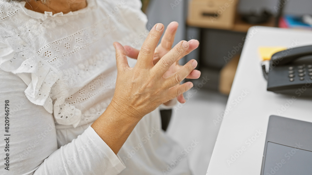 Mature woman experiencing hand pain at her office desk, perhaps from arthritis or overuse.