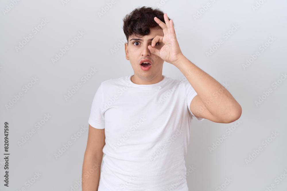 Young non binary man wearing casual white t shirt doing ok gesture shocked with surprised face, eye looking through fingers. unbelieving expression. Young non binary man wearing casual white t shirt doing ok gesture shocked with surprised face, eye looking through fingers. unbelieving expression.