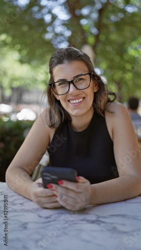 Smiling beautiful hispanic woman, cheerfully using smartphone while sitting at cafe's outdoor terrace, enjoying a coffee, connection with the digital world enhancing her happiness.