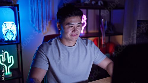 A young asian man focuses intently on his computer in a dimly lit room decorated with neon lights, reflecting a modern, tech-savvy lifestyle. © Krakenimages.com