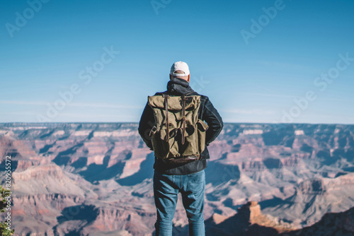Back view of male traveler enjoying view from mountain top getting to destination of hiking tour, hipster guy with backpack standing on rocky hill looking at dry valley in Grand canyon National Park