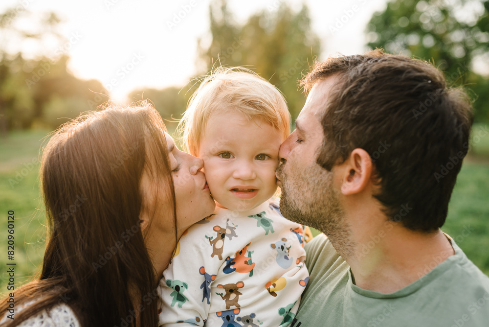 Parents kiss kid and happiness playing in green grass in park. Happy mother, father hug baby son walking in garden at sunset. Family spending time together outdoors. Children's day. Friendly family. Parents kiss kid and happiness playing in green grass in park. Happy mother, father hug baby son walking in garden at sunset. Family spending time together outdoors. Children's day. Friendly family.