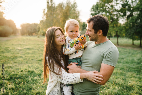 Happy mother, father hug baby son walking in garden at sunset. Family spending time together outdoors. Parents hugging kid and happiness playing in green grass in park. Children's day. Friendly family