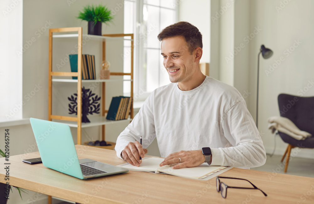 Portrait of young attractive business man working on a laptop at office or at home planning daily meeting and appointment looking cheerful at camera. Remote and freelance work concept.