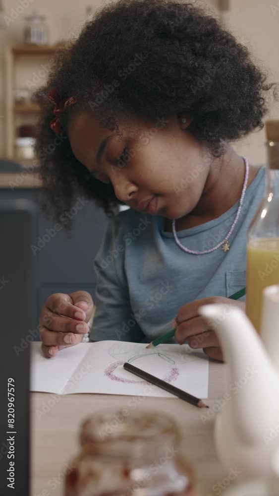 Medium vertical of young curly African American girl enthusiastically completing her drawing using colour pencils, sitting at table at home during daytime Medium vertical of young curly African American girl enthusiastically completing her drawing using colour pencils, sitting at table at home during daytime