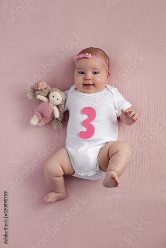 girl baby infant with a bow lies on the bed in a white bodysuit with a doll toy next to it, the child has a pink number 3 months on a pink background