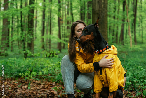 young woman walking with her doberman dog in the forest. Wearing yellow rain jackets girl and dog playing outdoors. Human and dog friendship concept
