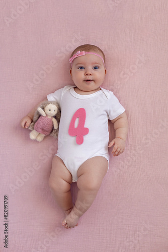 girl baby infant with a bow lies on the bed in a white bodysuit with a doll toy next to it, the child has a pink number four months on a pink background