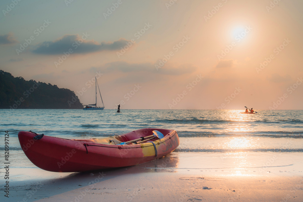 Tranquil beach scene at sunset with a red kayak on the shore, a sailboat, and kayakers in the distance. Tranquil beach scene at sunset with a red kayak on the shore, a sailboat, and kayakers in the distance.