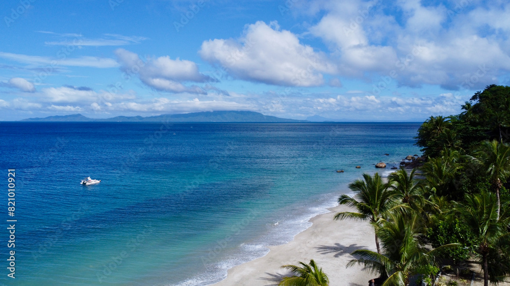 Aerial view of the coast of a tropical island. Top view of a sandy beach with palm trees, the surface of a calm sea, clouds in the sky and a small boat in the sea. Aerial view of the coast of a tropical island. Top view of a sandy beach with palm trees, the surface of a calm sea, clouds in the sky and a small boat in the sea.