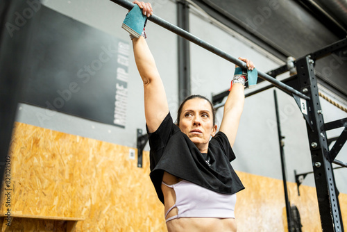 A middle-aged adult woman performs physical exercise inside a gym.The 50-year-old is hanging on a bar while looking sideways very concentrated. photo