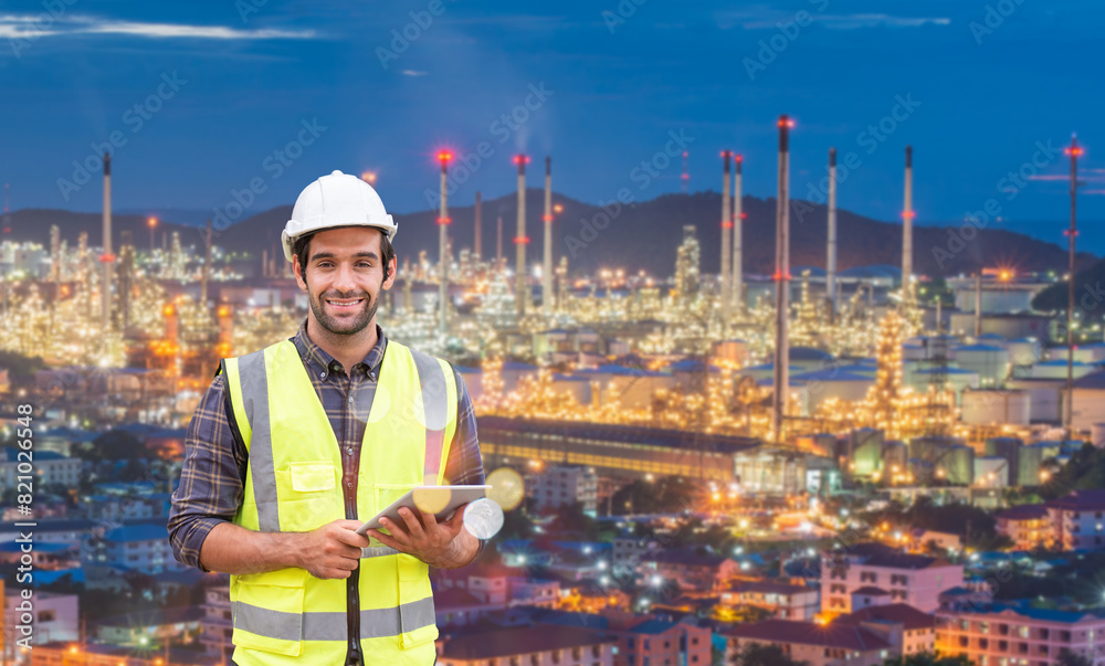 Smiling engineer with tablet and safety gear standing in front of illuminated industrial plant at night. Smiling engineer with tablet and safety gear standing in front of illuminated industrial plant at night.