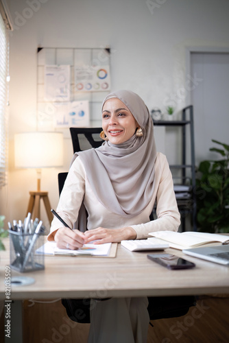 Smiling Muslim woman writing notes at office desk. Professional workspace, casual attire