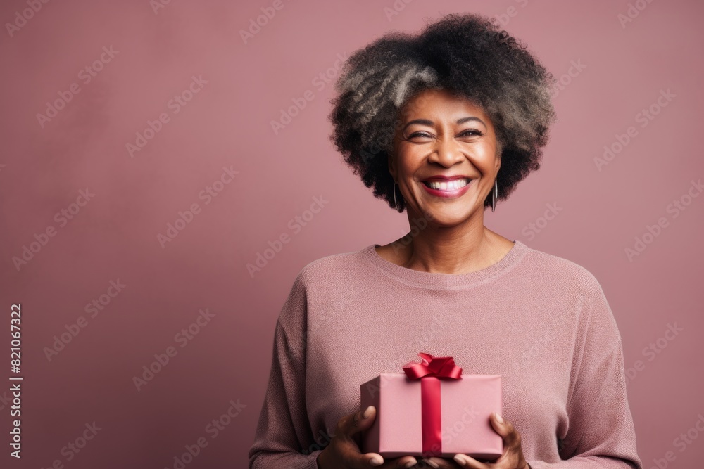 Portrait of a grinning afro-american woman in her 60s holding a gift while standing against minimalist or empty room background Portrait of a grinning afro-american woman in her 60s holding a gift while standing against minimalist or empty room background