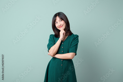 Thoughtful Asian woman keeps hand on chin looks pensively above dressed in green dress poses against green background blank copy space for your advertising content thinks about future