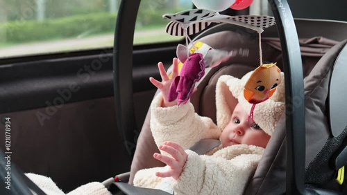 An infant lies in a car seat playing with a mobile, with a forest passing by in the background as the car moves. Close-up of the baby playing with the mobile