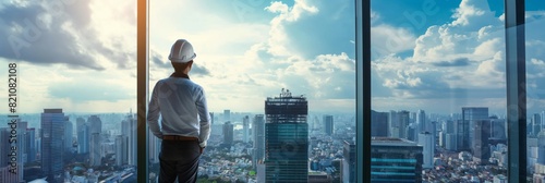 A male engineer in a hard hat stands contemplating the cityscape from a high vantage point in an office photo
