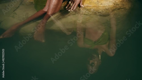 Reflection of androgynous black lgbtq fashion model lies on stones inside picturesque natural pool at night. Non-binary biethnic person poses in still water among rocks, touches water with hand photo