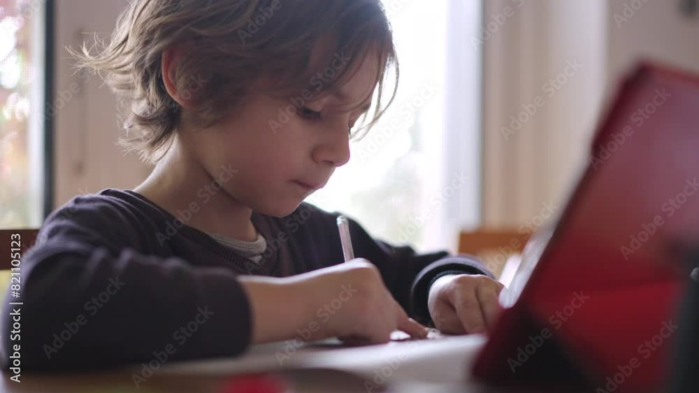 A focused young boy draws attentively on his digital tablet while sitting at a table in a well-lit room. A focused young boy draws attentively on his digital tablet while sitting at a table in a well-lit room.