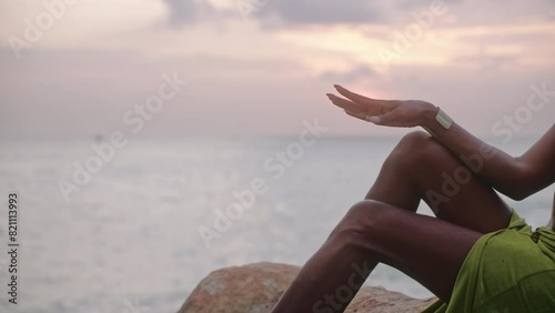 Trans sexual ethnic tender hand moves against sun with brass accessories in elegant posture sitting on stone in slow motion. Epatage gay black man poses above stormy ocean beach on sunset. photo