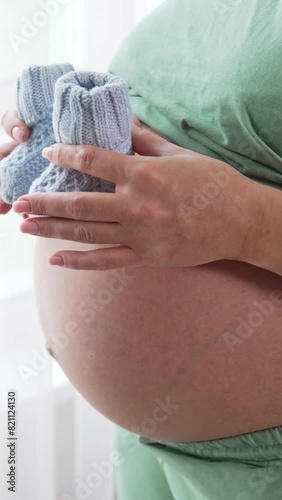 Unidentified pregnant woman paces small baby socks on the top of her big tummy and moves them slowly downwards. Future mom plays with knitted baby booties. Close up. Vertical video photo