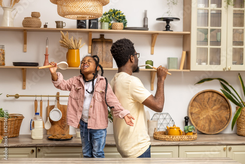Little adorable kid girl imagining artist singing favorite song with playful young father in kitchen