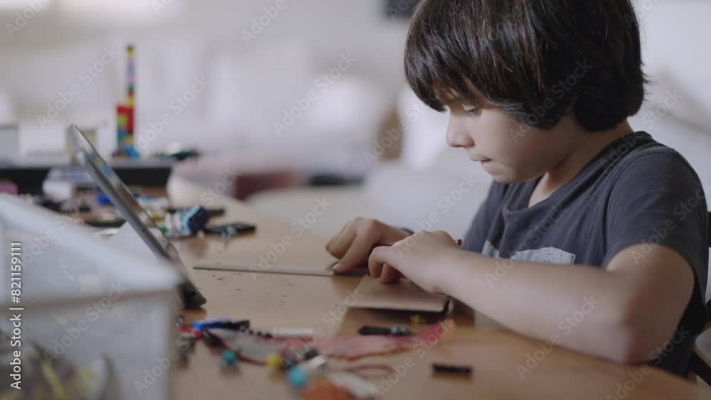 A young boy concentrates on a paper craft project while referring to instructions on a digital tablet at a table in a well-lit room. A young boy concentrates on a paper craft project while referring to instructions on a digital tablet at a table in a well-lit room.