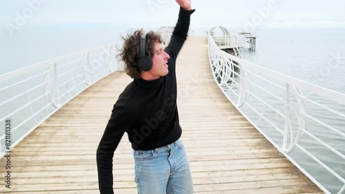 Cute young guy party-goer enjoying music in headphones, having fun dancing standing on the shore of the lake in happy solitude with his arms outstretched, celebrating acceptance to university.