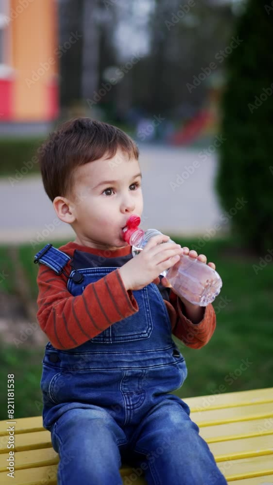 Lovely toddler boy sits on the bench holding a water bottle in hands. Kid drinks water outdoors. Soap bubbles at foreground. Vertical video. Lovely toddler boy sits on the bench holding a water bottle in hands. Kid drinks water outdoors. Soap bubbles at foreground. Vertical video.