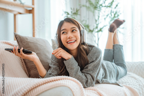 Happy asian woman listening to music from mobile phone while sitting on the the sofa at homes, Smiling girl relaxing with headphones in morning, Time to relax. © oatawa