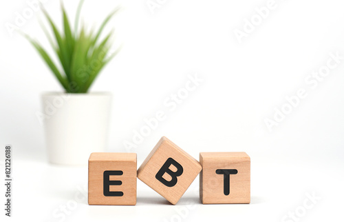 3 wooden cubes with letters on table with a pot of green plant on white background. The words written is EBT Earnings Before Taxes acronym. Business Concepts. Idea of finance.Front View, Copy space.