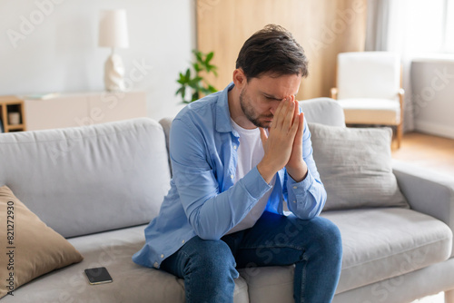 A young man is seated on a grey sofa, looking distressed with his hands covering his face. A smartphone lies nearby, suggesting he may have received upsetting news © Prostock-studio