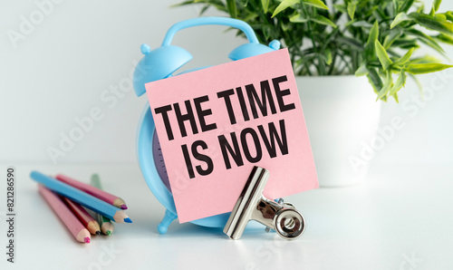 Paper note word The Time Is Now on alarm clock desk white background. Sticker note paper message Now and watch on wooden table.