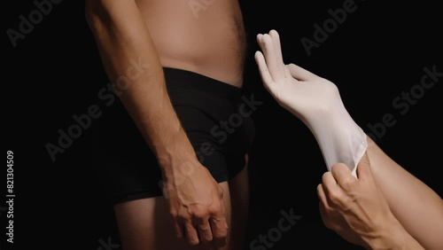 A Female Doctor Puts on Gloves Before Examining a Man in Underwear Standing in Front of Her, Reaching Toward His Genitals. Testicular Examination.