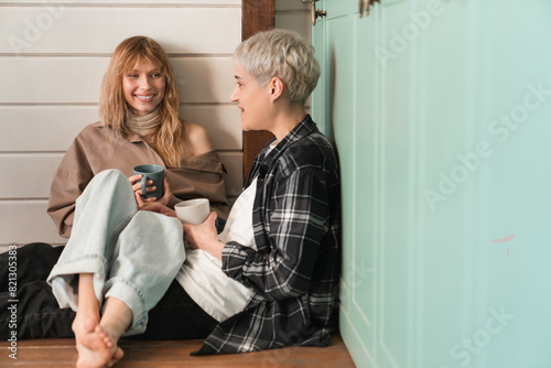 Fototapeta Young caucasian female LGBT lovers, young girlfriends talking on the kitchen floor drinking coffee or tea together showing their passion and love