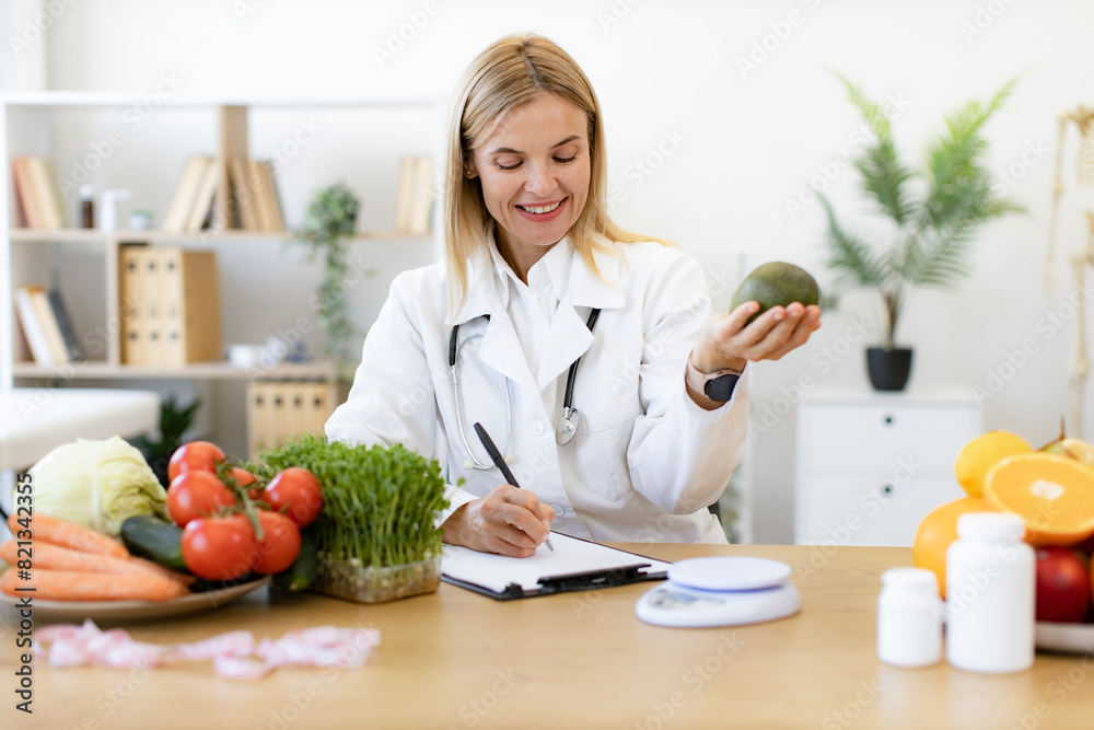 Happy female writing prescription for patients proper healthy diet. Mature Caucasian female nutritionist making healthy eating plan and calculating calorie content of avocado using weight. Happy female writing prescription for patients proper healthy diet. Mature Caucasian female nutritionist making healthy eating plan and calculating calorie content of avocado using weight.