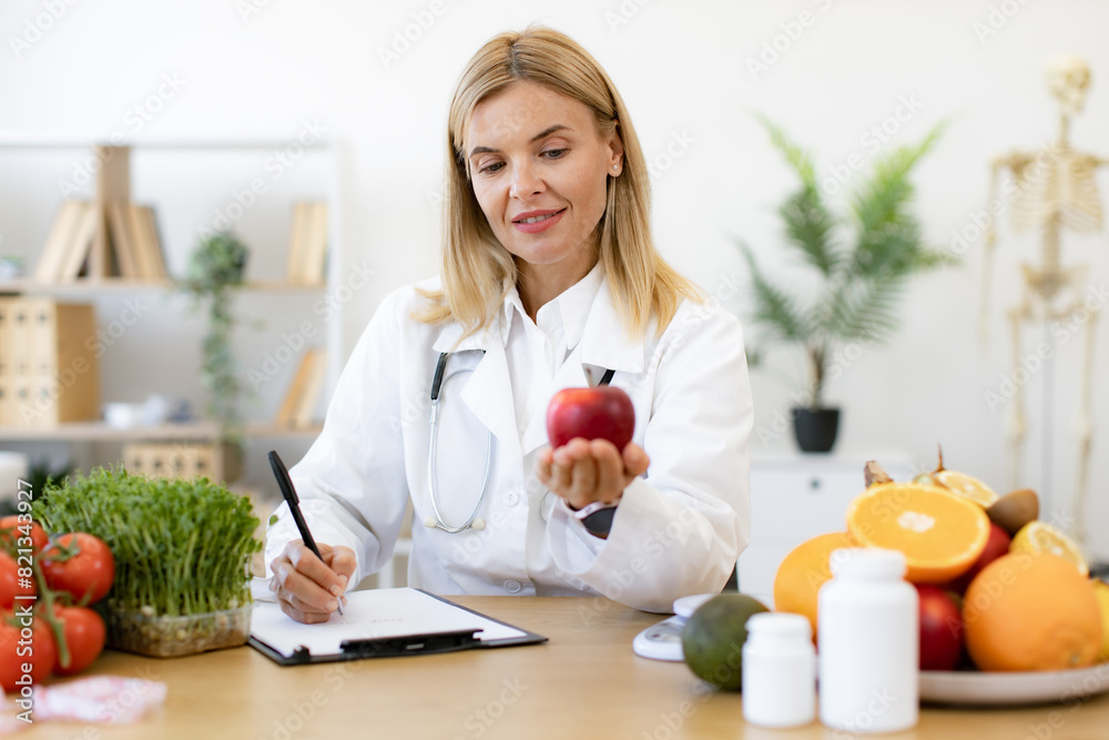 Portrait of smiling female posing with apple while doing paperwork in doctor's office. Caucasian nutritionist woman including client's preferences about fruits into meal plan. Portrait of smiling female posing with apple while doing paperwork in doctor's office. Caucasian nutritionist woman including client's preferences about fruits into meal plan.