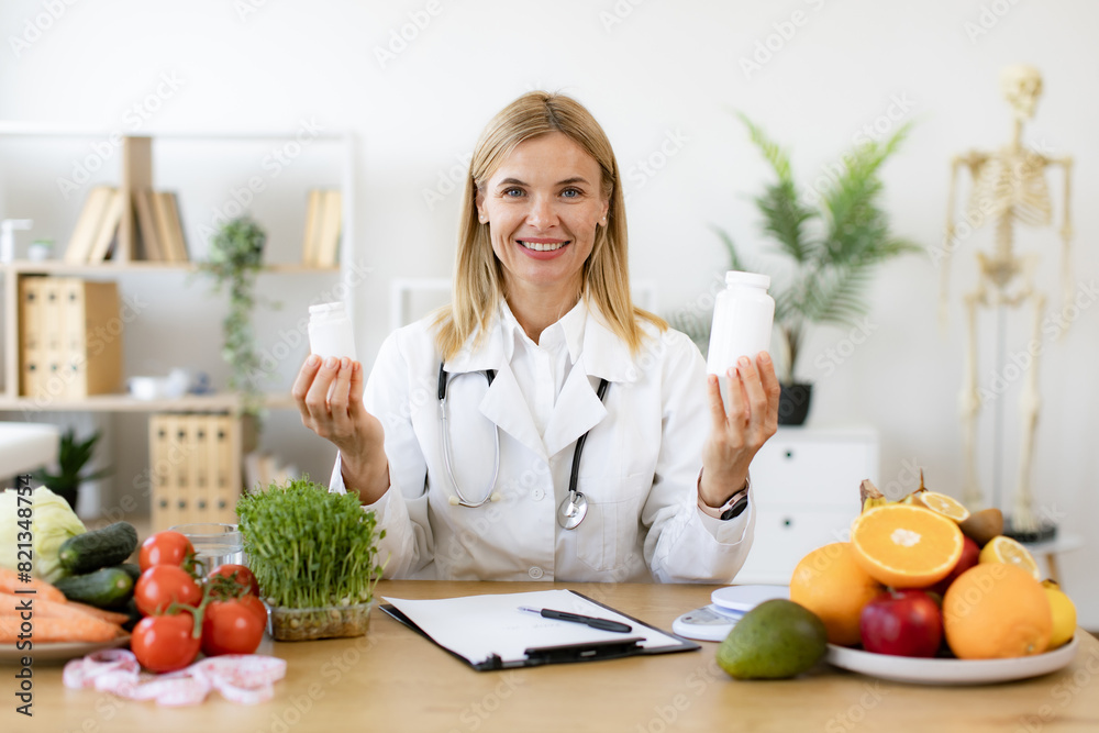 Portrait of positive Caucasian lady in lab coat smiling at camera while demonstrating two pills bottles in office. Cheerful expert in food proposing dietary supplements to improve healthy meals. Portrait of positive Caucasian lady in lab coat smiling at camera while demonstrating two pills bottles in office. Cheerful expert in food proposing dietary supplements to improve healthy meals.