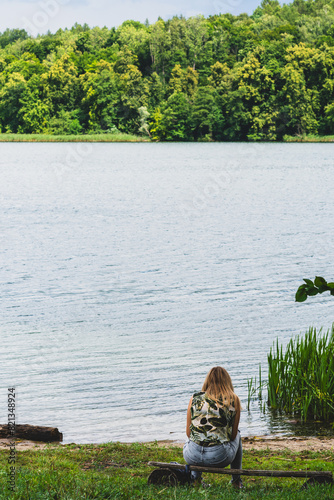 A girl sits on a wooden bench by the lake in warm summer weather. Thinking one on one with nature.