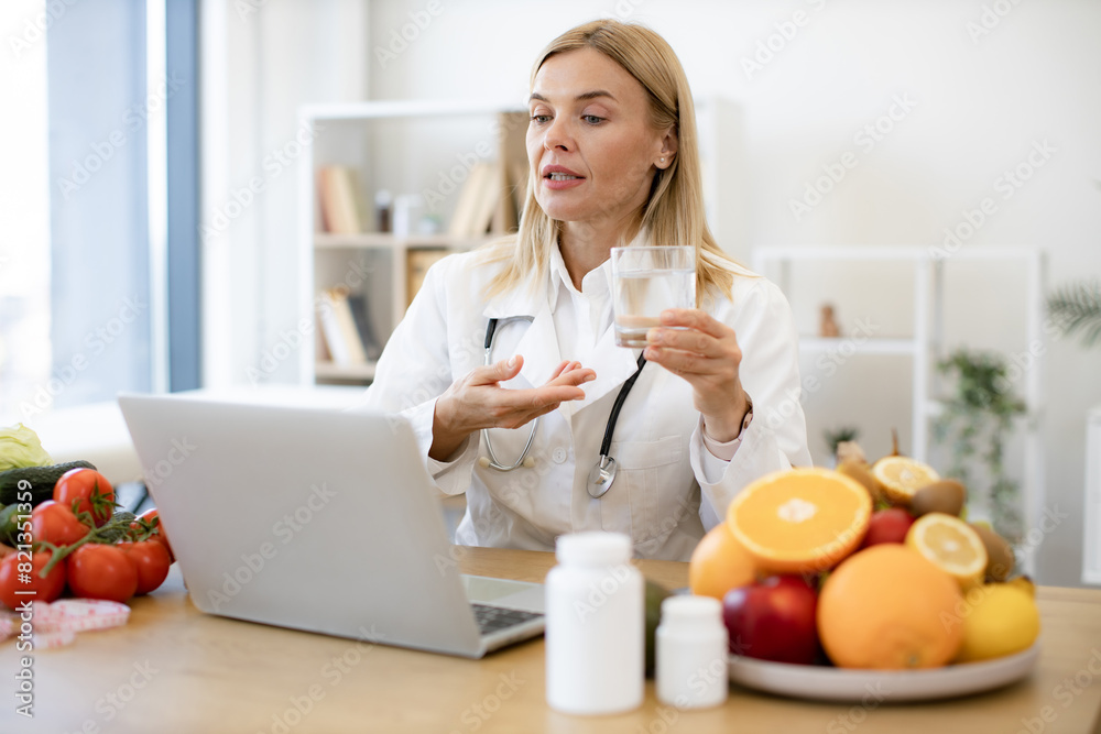 Female doctor in white coat holding glass of water during a video call. Experienced female nutritionist tells patient about benefits of water during an online session using laptop. Female doctor in white coat holding glass of water during a video call. Experienced female nutritionist tells patient about benefits of water during an online session using laptop.