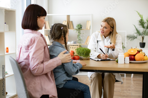 Mature Caucasian female doctor talking with child girl sitting on her mom knees and writing diet plan. Experienced female dietitian prepares course of healthy nutrition for small child.
