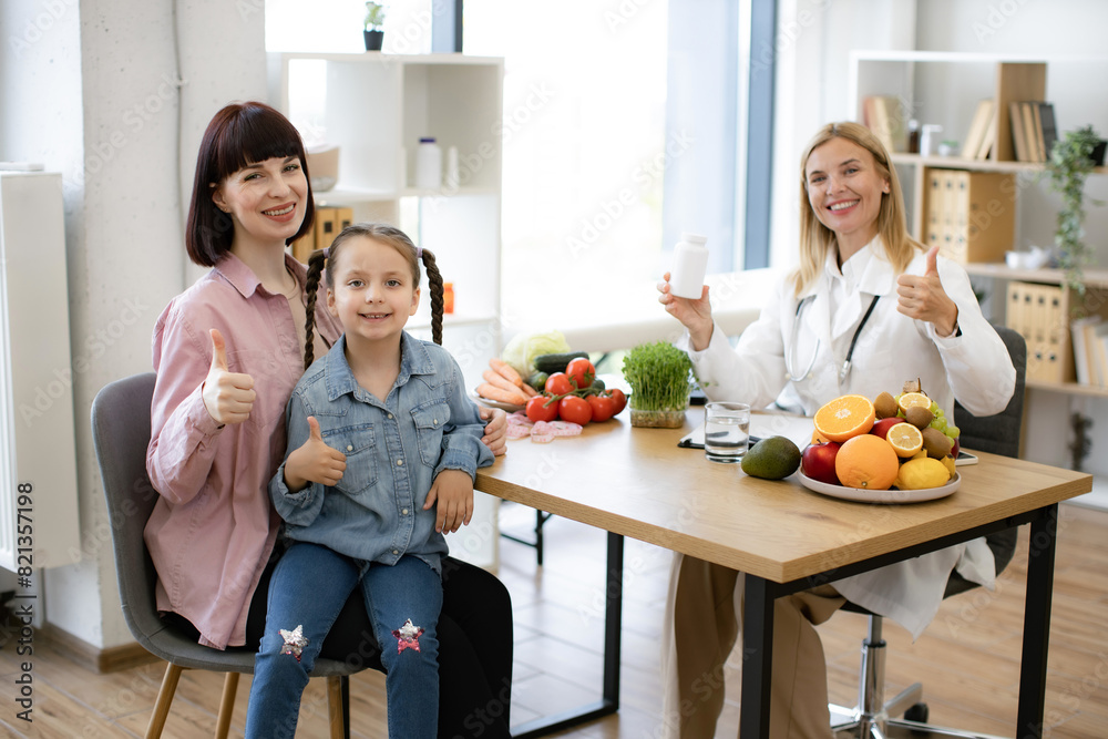 Portrait of female nutritionist holding bottles with pills as nutrition supplements for little girl sitting on moms lap, looking at camera. Mature pediatrician receives female patient in modern clinic Portrait of female nutritionist holding bottles with pills as nutrition supplements for little girl sitting on moms lap, looking at camera. Mature pediatrician receives female patient in modern clinic