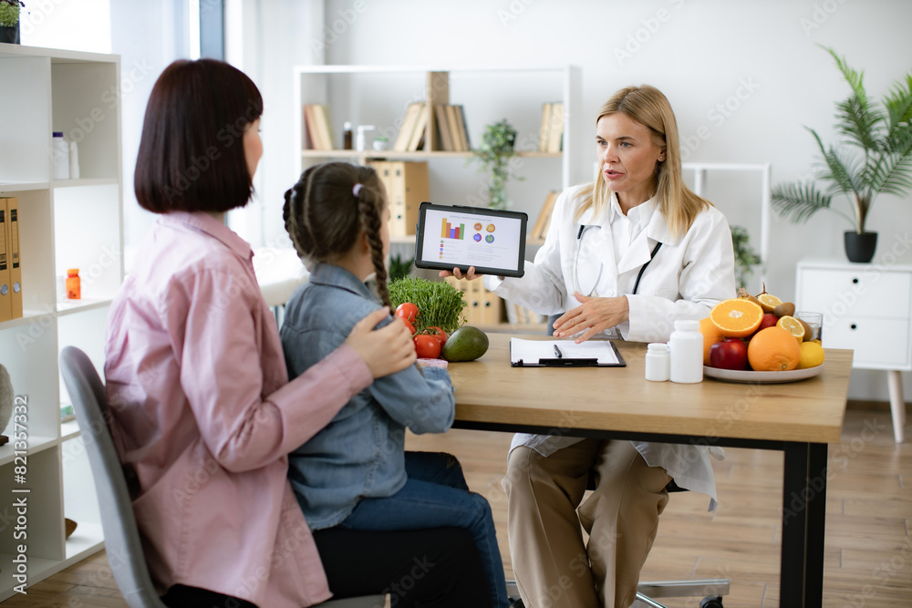 Fototapeta premium Little school girl sitting on mothers lap with visit to woman pediatrician nutritionist. Mature Caucasian female doctor shows on tablet graphs of food for healthy eating.