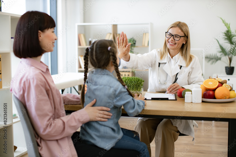 Little girl sits on mothers lap and gives high five to mature positive female nutritionist after successful treatment for indigestion. Concept of completing gastrointestinal treatment plan. Little girl sits on mothers lap and gives high five to mature positive female nutritionist after successful treatment for indigestion. Concept of completing gastrointestinal treatment plan.