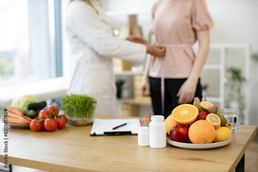 Close up view of woman in doctor's coat finding out waist length of female patient with measuring tape in consulting room. Efficient dietitian calculating body mass index for assessing healthy weight. Close up view of woman in doctor's coat finding out waist length of female patient with measuring tape in consulting room. Efficient dietitian calculating body mass index for assessing healthy weight.
