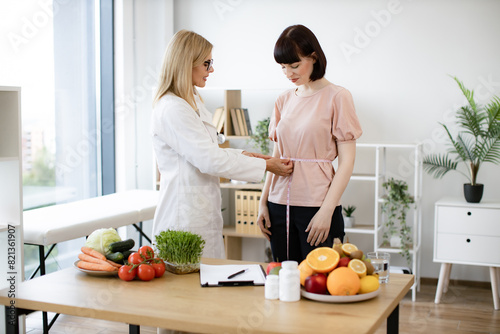 Mature woman in doctor's coat finding out waist length of female patient with measuring tape in consulting room. Efficient dietitian calculating body mass index for assessing healthy weight. © sofiko14