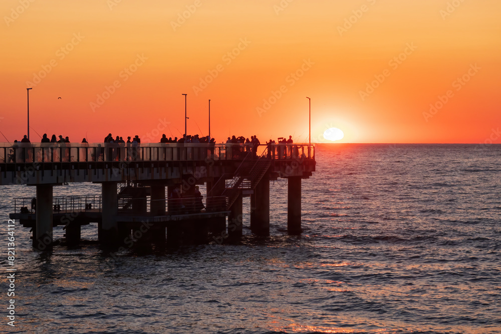 Many people watch the sunset from the pier over the sea. The orange sun was half hidden behind the horizon. Background.