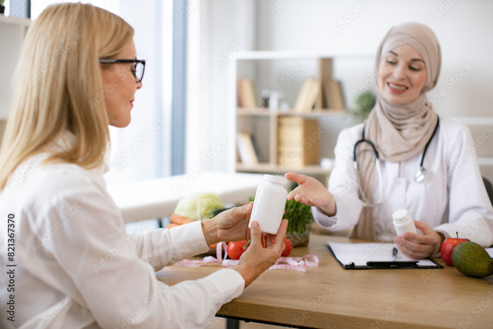 Obraz premium Mature Caucasian lady receives consultation on healthy eating. Muslim woman nutritionist makes plan for treatment of gastrointestinal tract using vitamins and supplements for healthy diet.