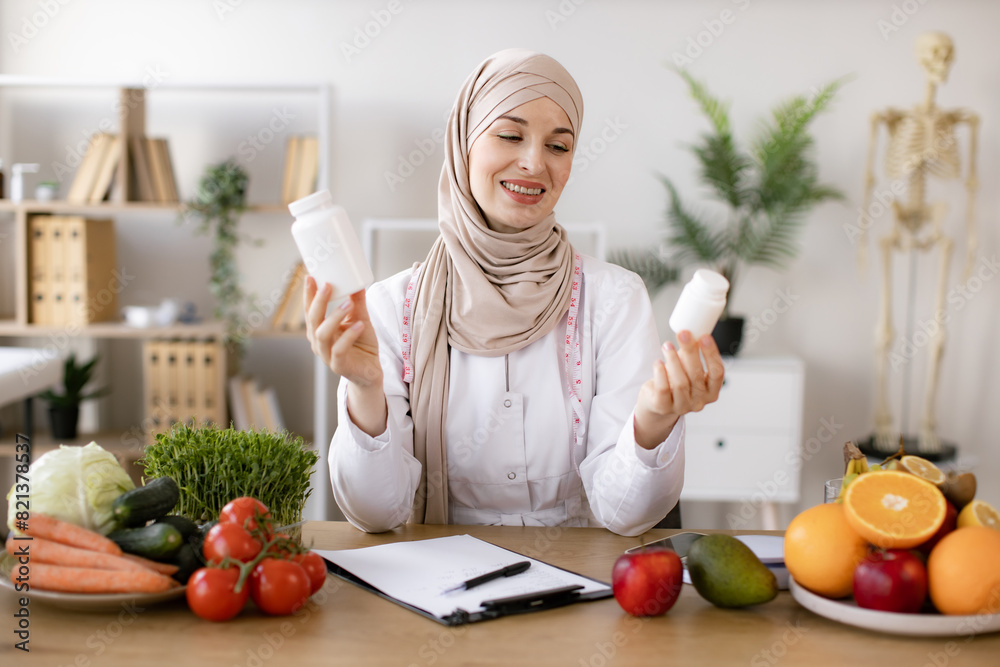 Cheerful expert in food proposing dietary supplements to improve healthy meals. Portrait of positive Muslim lady in hijab and lab coat smiling at camera while demonstrating two pills bottles in office Cheerful expert in food proposing dietary supplements to improve healthy meals. Portrait of positive Muslim lady in hijab and lab coat smiling at camera while demonstrating two pills bottles in office
