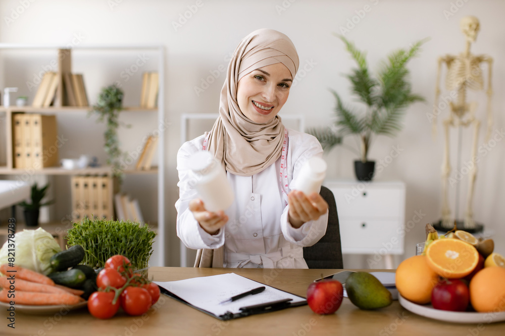 Portrait of positive Muslim lady in hijab and lab coat smiling at camera while demonstrating two pills bottles in office. Cheerful expert in food proposing dietary supplements to improve healthy meals Portrait of positive Muslim lady in hijab and lab coat smiling at camera while demonstrating two pills bottles in office. Cheerful expert in food proposing dietary supplements to improve healthy meals